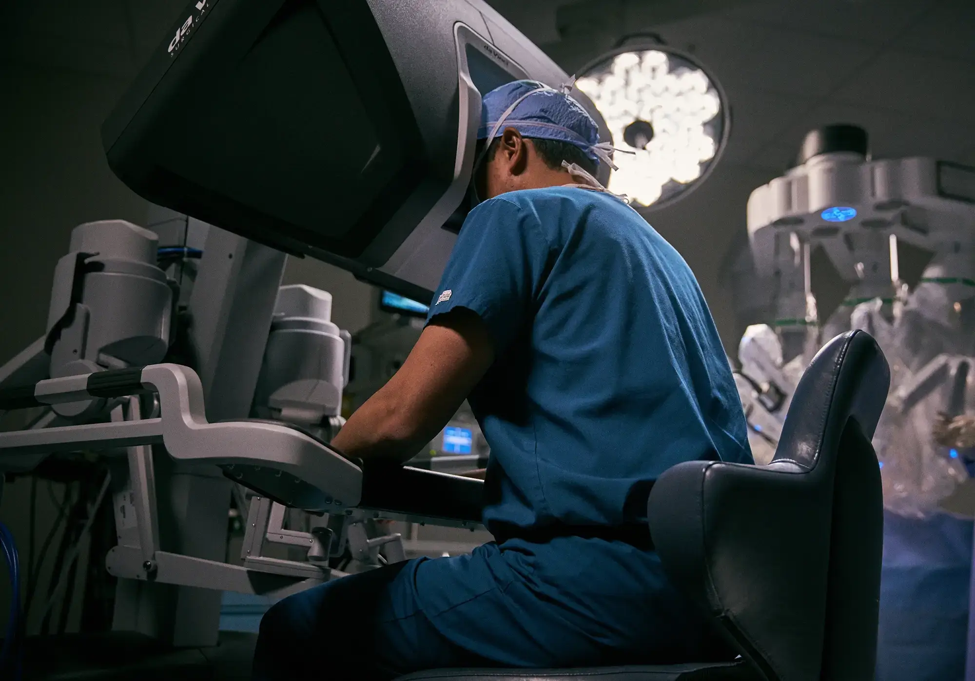 Dr. Wei Phin Tan performing robotic prostatectomy at the da Vinci surgical console at NYU Langone — blue scrubs and surgical cap, operating room lights and robotic arms visible